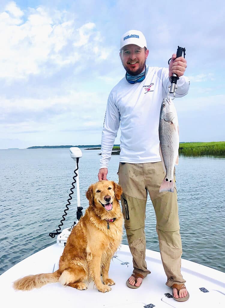 A man holds a fish aboard a boat with his dog at his side